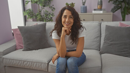 A smiling middle-aged brunette woman sitting comfortably on a couch in a modern living room interior
