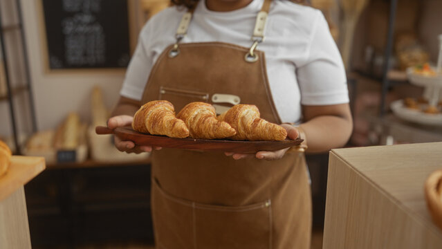 Young african american woman in a bakery holds a tray of croissants, dressed in an apron over a white shirt, indoor scene showcasing freshly baked pastries. - Powered by Adobe