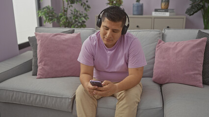 Obraz premium A young hispanic man listens to music on his smartphone while relaxing on a sofa in a modern apartment living room.
