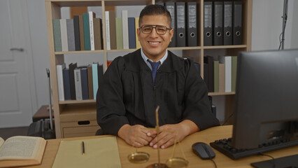 Young hispanic man in black robe sitting at a desk in an office with a computer and legal books,...