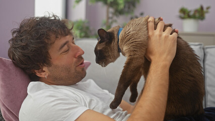 A young man affectionately pets his siamese cat in a cozy living room, creating an image of comfortable domestic life.