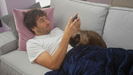 A young hispanic man lounges on a sofa at home, engrossed in his smartphone with a siamese cat...