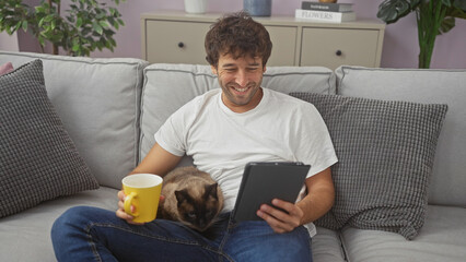 A smiling young man with a coffee mug enjoying a tablet with his siamese cat on a sofa indoors.