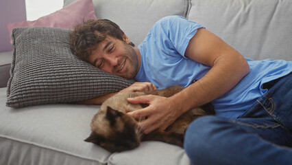 A smiling man cuddles with a siamese cat on a couch in a cozy living room setting