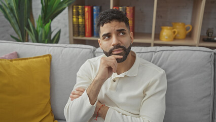 A thoughtful hispanic man sits on a gray couch in a cozy living room, conveying introspection.