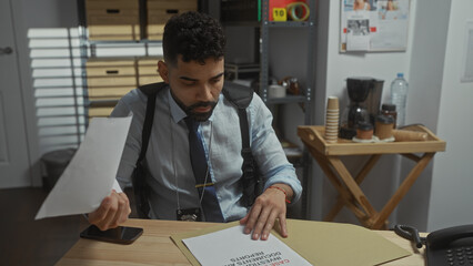 A focused hispanic man with a beard examines documents at a desk in a detective's office.