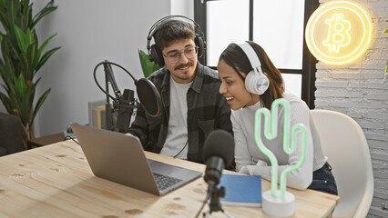 Man and woman wearing headphones smiling while recording a podcast in a studio decorated with a neon bitcoin sign.