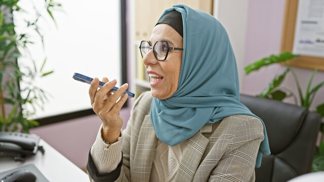 Smiling woman wearing hijab and glasses holding smartphone in modern office setting.