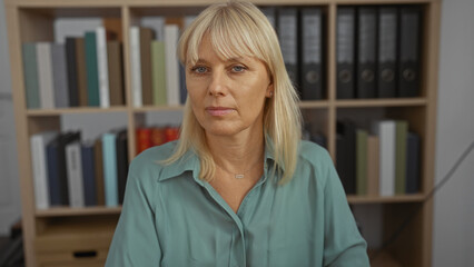 Mature woman with blonde hair sitting in an office with a bookshelf full of binders and books in...