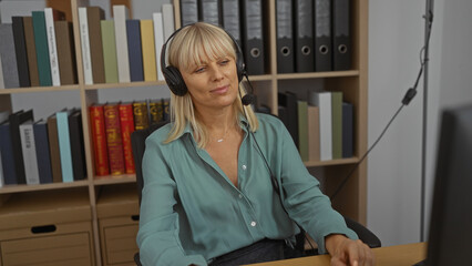 Woman working in an office wearing a headset, sitting at a desk with bookshelves in the background...