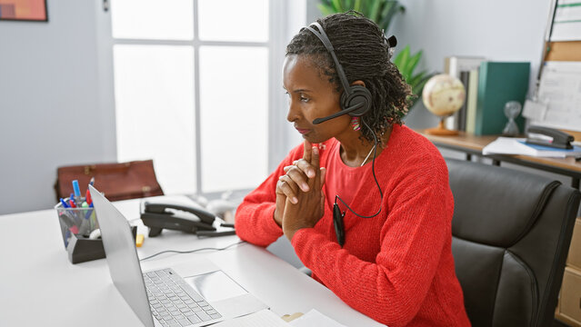 African american woman using a laptop and headset in a modern office setting, focused and professional.