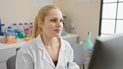 A young blonde woman in a laboratory wearing a white coat sitting at a desk, looking thoughtful.