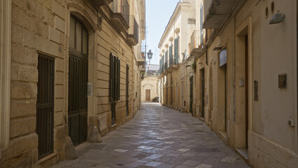 A quiet, narrow street in lecce, puglia, italy, showcasing historic architecture and mediterranean charm with closed shutters and cobblestone paving on a sunny day.