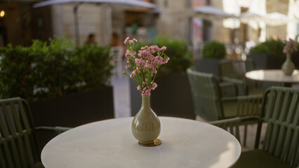 A beautifully set outdoor cafe table in a street of puglia, italy, showcasing a delicate bouquet of pink italian statice flowers in a vase with cozy chairs and lush green plants in the background.