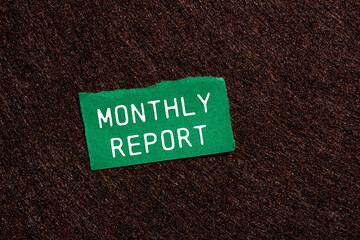 Monthly report message written on ripped torn green paper piece on brown background. Conceptual...