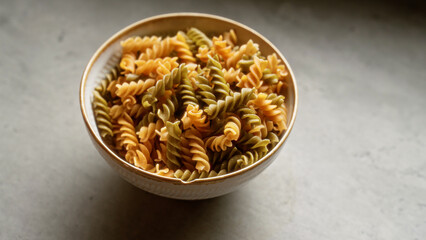 Full bowl of uncooked fusilli pasta in mixed colors resting on a smooth gray surface in soft natural light, showcasing food ingredients for cooking or recipe preparation