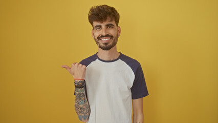 Handsome young man with beard smiling and pointing left over isolated yellow background