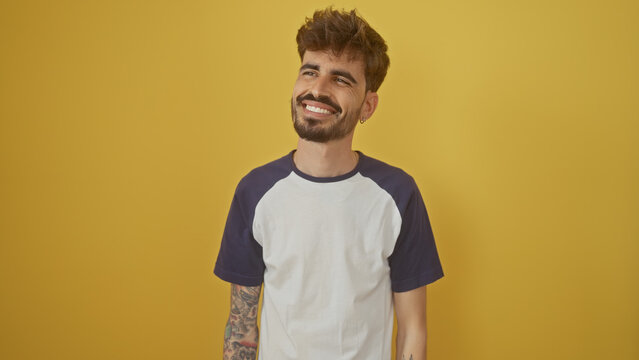 A handsome young hispanic man with a beard and tattoos smiles casually against a vibrant yellow background.