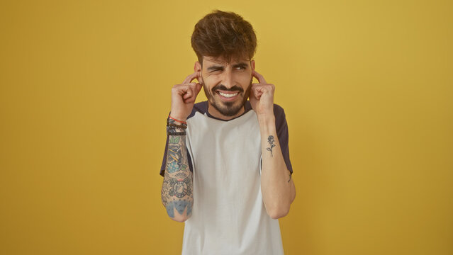 A young hispanic man with a beard and tattoos smiles against a yellow wall, covering his ears with his hands.