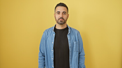 Handsome hispanic man with beard in denim jacket against yellow background