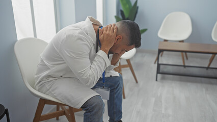 A stressed hispanic man in a white coat holding his head in a hospital waiting room