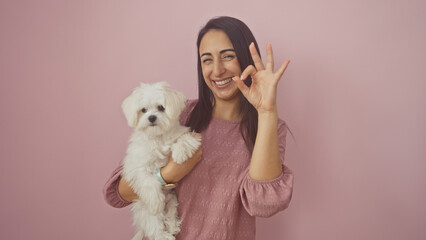 A happy young adult woman holding a white maltese dog, making an ok hand sign against a pink...