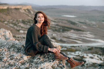 Confident woman in sweater and boots sitting on mountaintop with hands on hips overlooking scenic view