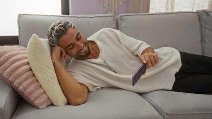 Obraz premium Young man with beard watching video on mobile while lying on couch in cozy living room