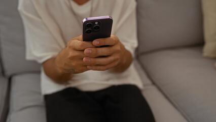 Young man holding smartphone while sitting indoors on a couch in a living room, focused on his hands and phone, wearing casual clothing in a modern apartment setting.