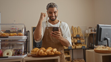 Young man with beard in bakery wearing apron looking at smartphone celebrating success surrounded by bread and pastries