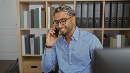 Young man with beard talking on smartphone in an office workspace filled with bookshelves and...