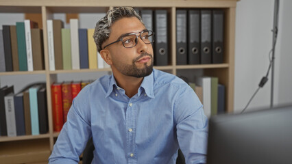 Young, arab, muslim man with a beard sitting in an office workspace indoors, surrounded by shelves filled with books and folders