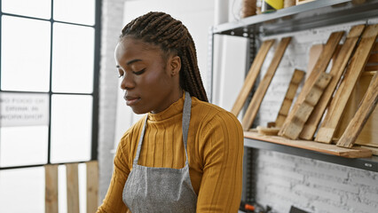 A contemplative african american woman with braids wearing an apron stands in a carpentry workshop.