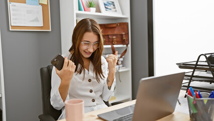 Young brunette woman celebrates success in modern office with laptop and smartphone.