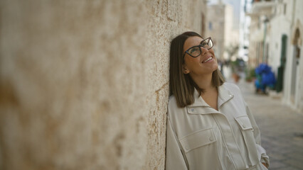 Beautiful young hispanic woman smiling in the charming streets of polignano a mare, puglia, italy, leaning against a stone wall on a sunny day.