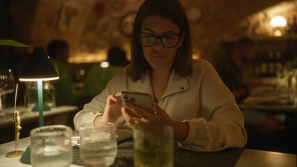 A young, beautiful hispanic woman sits indoors at an italian restaurant, engaging with her phone and surrounded by glasses on the table.