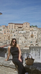 A beautiful young hispanic woman sits at a viewpoint in the old town of matera, italy, with a charming backdrop of historic buildings.