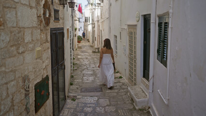 A young beautiful hispanic woman in a white dress strolls through the charming narrow streets of ostuni old town in puglia, italy, europe.