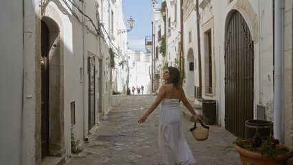 Young hispanic woman in white dress exploring charming old town of ostuni, italy, with narrow cobblestone streets and traditional buildings.