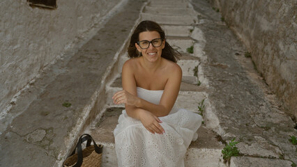 A beautiful young hispanic woman in a white dress sits on rustic stone steps in ostuni, an old town in puglia, italy, smiling warmly with a wicker bag beside her.