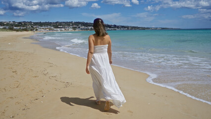Young hispanic woman in a white dress walks along the sandy beach of pescoluse in puglia, italy, enjoying the scenic salento coastline under a bright blue sky.