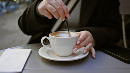 Close-up of a woman stirring coffee on a terrace table with smartphone beside.