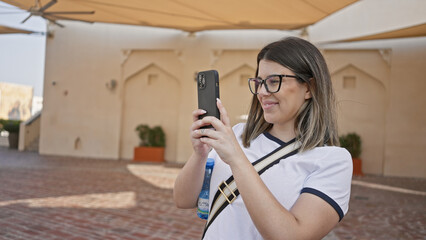 Smiling woman using smartphone in doha, capturing traditional arabic architecture on a sunny day.