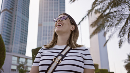 Fototapeta premium Happy woman laughing against abu dhabi skyline with modern skyscrapers, palms, and sunny urban settings.