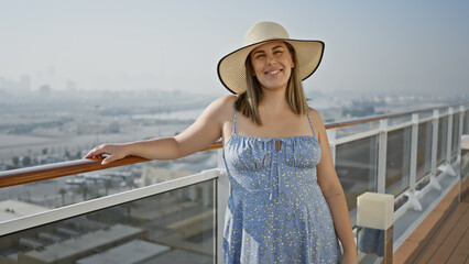 A smiling young woman in a sundress and hat leans on a cruise ship's railing, gazing out at the sea.