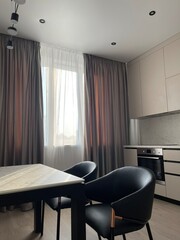 Completed modern repair, the sun shines through the gray curtain and tulle. Black leather chairs with copper handles are standing at the marble table. 