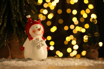 Cute decorative snowman on artificial snow against blurred lights. Space for text