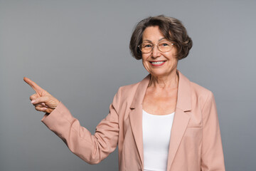 Mature senior smiling businesswoman wearing glasses in smart casual standing over isolated grey background cheerful pointing with hand and finger up to the side. Advertisement concept