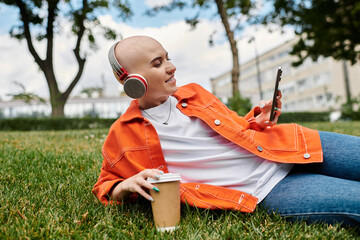 A bald woman relaxes on the grass, listening to music while sipping her coffee.