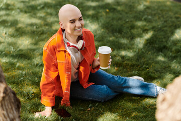 A cheerful young woman with alopecia sits on the grass sipping coffee, basking in sunlight.